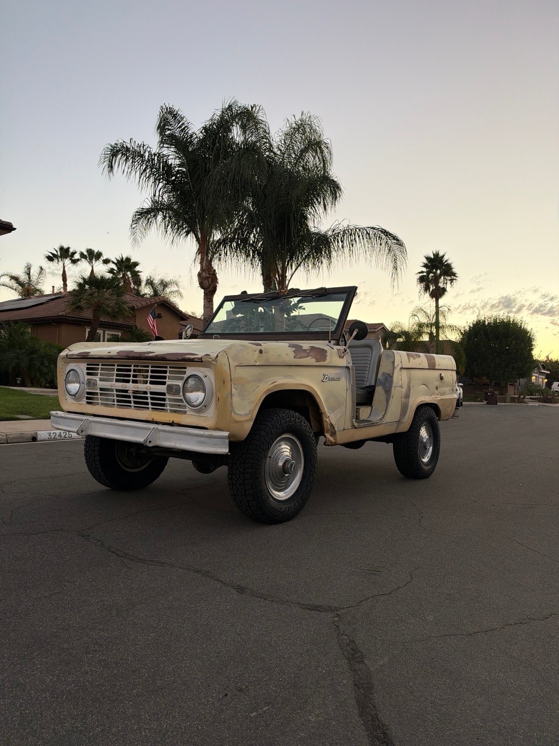 The pale yellow 1966 Ford Bronco U13 survivor, with weathered paint and an open cabin, beneath palm trees at sunset.